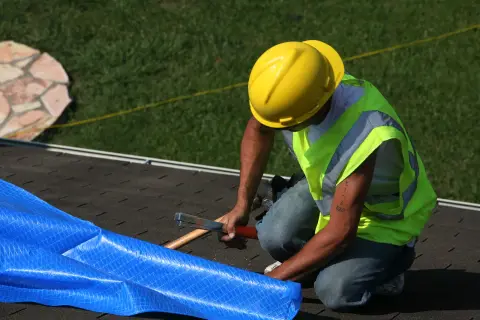 Worker climbing on a residential roof to secure a blue emergency tarp over storm damage