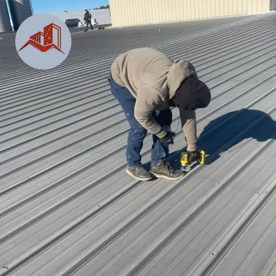 Roofer fastening ribbed gray metal roofing panels on a commercial low-slope roof with a cordless drill